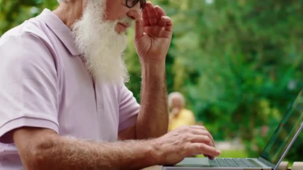 Beau vieil homme avec une barbe très fatigué enlever des lunettes de soleil après qu'il a terminé son travail sur l'ordinateur portable au milieu du parc au café. Tourné sur ARRI Alexa Mini. — Video