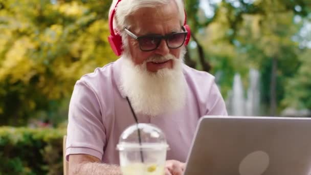 Před kamerou ve slunci uprostřed parku poslouchá hudbu ze sluchátek a pracuje na laptopu a užívá si čas na čerstvém vzduchu — Stock video