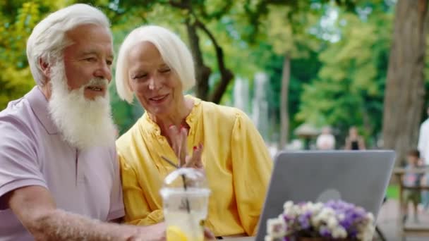 Посреди парка в кафе очень красивая пожилая пара проводит время в чате с ноутбука, обсуждая с кем-то, с кем они счастливо улыбаются — стоковое видео