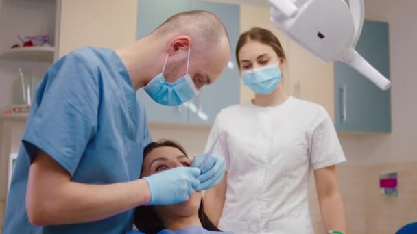 Dentista homem concentrado verificar os dentes de sua senhora paciente e seu assistente de pé ao lado do dentista — Vídeo de Stock