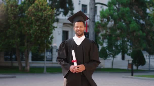 Όμορφος τύπος στην ημέρα αποφοίτησής του φορώντας κοστούμι και καπέλο αποφοίτησης και ποζάροντας μπροστά στην κάμερα με το δίπλωμα του στον κήπο του κολεγίου — Αρχείο Βίντεο