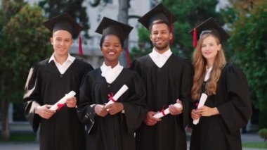 Bir grup çok ırklı öğrenci, diplomalarıyla kameranın önünde poz vererek mezun oldular ve üniversite bahçesinde mezuniyet kıyafeti ve şapkaları içinde heyecanlandılar.