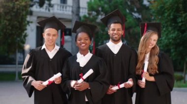 Üniversite bahçesinde, kameranın önünde çok ırklı mezunları poz vererek kameraya bakıp gülümseyen erkekler ve bayanlar.
