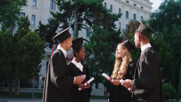 Podekscytowana grupa uczniów wielorasowych w dniu ukończenia szkoły rozmawiają ze sobą czując się szczęśliwi trzymając w rękach dyplomy — Wideo stockowe