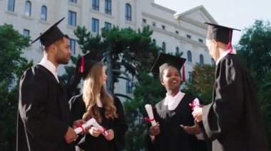 Üniversite bahçesinde mutlu ve gülümseyen bir grup çok ırklı öğrenci mezuniyet günlerinde diplomalarını ellerinin arasında tutarak anın tadını çıkarıyorlar.