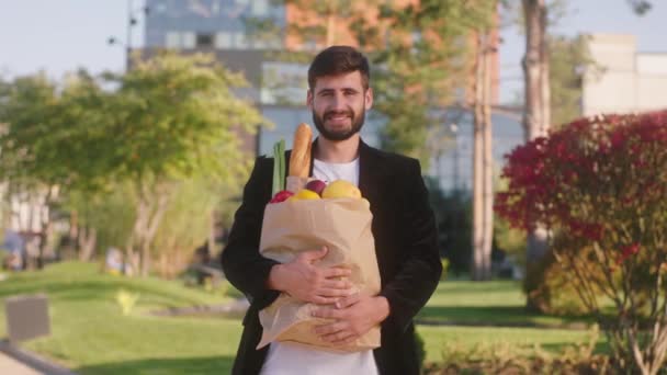 Перед камерой, изображающей харизматичного мужчину с широкой улыбкой посреди улицы, он держит эко-сумку, полную овощей и хлеба — стоковое видео