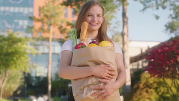 Caddenin ortasında alışveriş yaptıktan sonra gülümseyen güzel kadın kameranın önünde poz veriyor. Elinde sebze ve meyve dolu bir çanta tutuyor. — Stok video