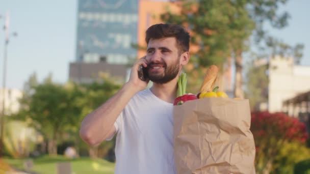 Perfektní slunečný den atraktivní muž kráčí po ulici s eko taškou plnou jídla mluví na svém smartphonu a vychutnat si čas — Stock video