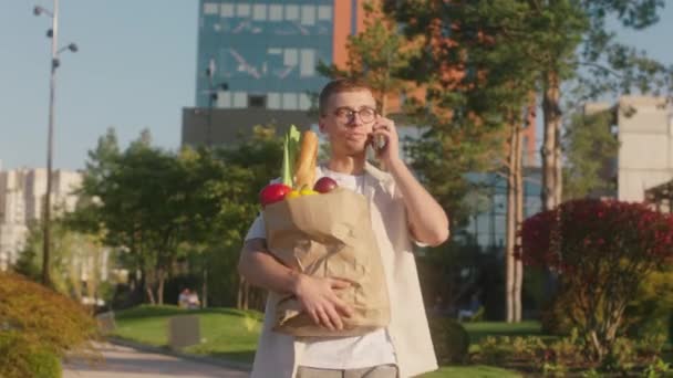 Przed kamerą młody mężczyzna przystojny po sklepie idący ulicą z eko torbą pełną warzyw i owoców i mówiący na smartfonie — Wideo stockowe