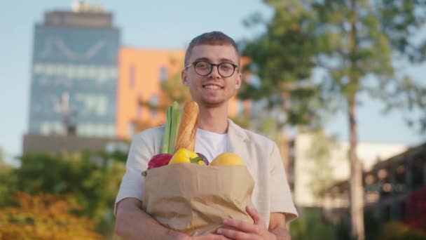 Posing seorang pria dengan kacamata di depan kamera di tengah kota modern ia memegang tas eco penuh sayuran dan buah-buahan — Stok Video