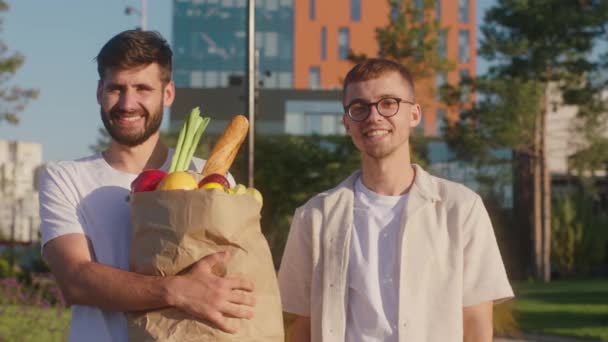 通りの真ん中にカメラの前にポーズをとっているカリスマ的な男2人の完璧な晴れた日には、野菜や果物でいっぱいのエコショップバッグを持っている男の1人 — ストック動画