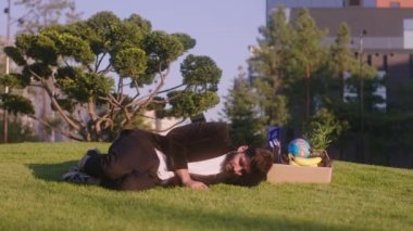 Modern bir parkta çimlerin üzerinde çok yorgun ve yakışıklı bir adam işten kovduğu eşyalarının üzerinde kestiriyor.
