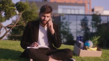 Güzel görünümlü genç adam ofis müdürü günün sonunda sokağın aşağısındaki modern parkta oturuyor birilerini aramaya başlıyor ve not defterine bazı notlar yazıyor.