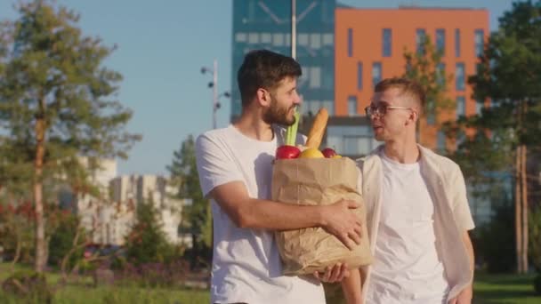 Solrig dag i midten af moderne gade gå sammen to smilende store fyre en af dem holder en øko taske med grøntsager og frugter. 4k – Stock-video