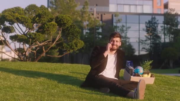 Перед камерою харизматичний хлопець лежить на траві насолоджуватися сонцем, який він закінчив день роботи взяти свою коробку з особистими речами і говорить по телефону — стокове відео
