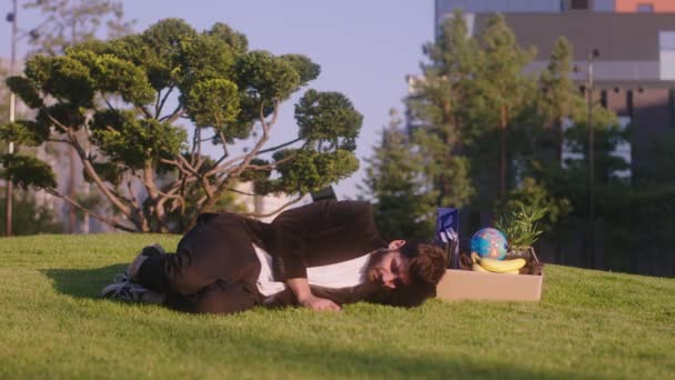 У сучасному парку вниз на траві красивий чоловік дуже втомився від своїх речей, які він звільнив з роботи — стокове відео