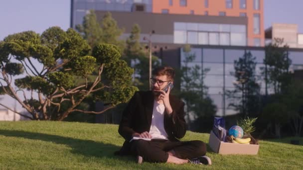 Em um moderno parque de negócios jovem trabalhador de escritório depois que ele terminar a semana de trabalho, sente-se na grama e começar a chamar alguém e fazer algumas notas no caderno — Vídeo de Stock