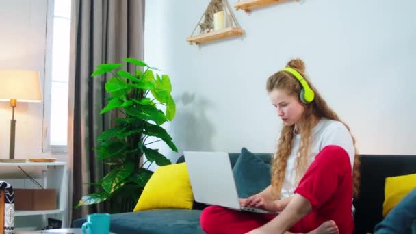 Auf dem Sofa im Wohnzimmer gut aussehende Frau konzentriert arbeiten vom Laptop mit Kopfhörern tippt sie einige Dokumente — Stockvideo