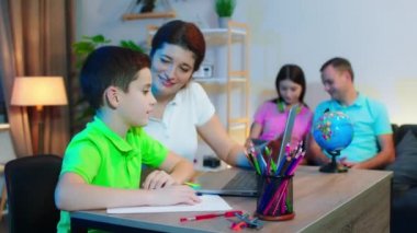 Modern geniş oturma odasında küçük sevimli bir çocuk ve annesi birlikte okul projesi yaparken diğer aile üyeleri kanepede oturup dijital tabletten bir şeyler izliyorlar.