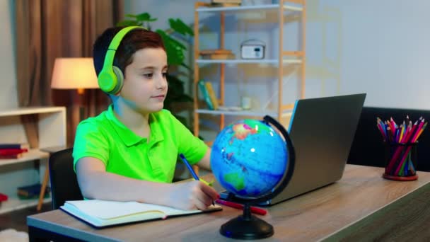 Na sala de estar concentrado menino bonito fazendo suas aulas on-line usando laptop e usando fones de ouvido verdes ele faz algumas notas no papel — Vídeo de Stock