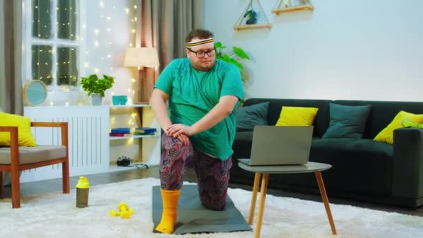 There 's a young obese man with a beard and black glasses, he is trying to do lunges but is struggling with them, he is wearing hippie pants and is on a workout mat, he is staring at his laptop while — стоковое видео