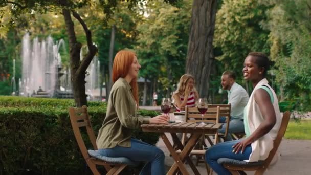 Un par de bellas mujeres guapas están bebiendo un poco de vino en una mesa en el hermoso parque, mientras que una pareja se sienta en una mesa detrás de ellas — Vídeos de Stock