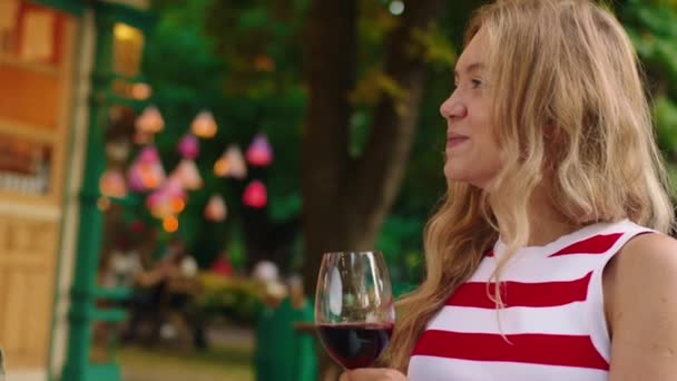 Een close-up shot van een prachtige jonge blonde vrouw, pratend en lachend terwijl ze een wijnglas vasthoudt, draagt ze een klassiek rood-wit gestreept vest — Stockvideo