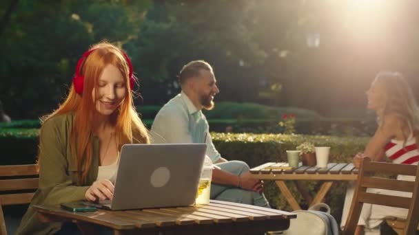 Μια νεαρή γυναίκα με ξανθά μαλλιά φοράει κόκκινα ασύρματα ακουστικά και κάνει δουλειά στο λάπτοπ της σε ένα τραπέζι σε δημόσιο κήπο, ενώ ένα ζευγάρι στο παρασκήνιο κάθεται σε ένα τραπέζι και μιλάει — Αρχείο Βίντεο