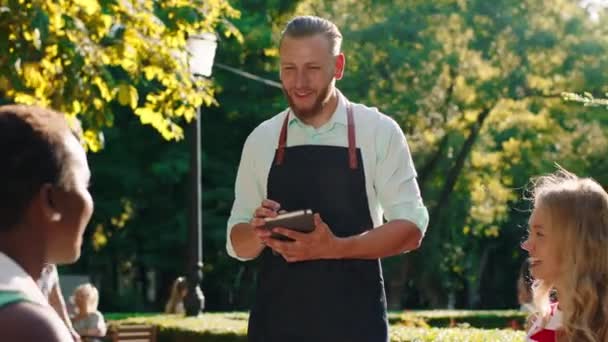 Bardzo przystojny kelner z zaczesanymi włosami, wykonuje rozkazy w swoim notatniku, rozmawia z trzema kobietami przyjmującymi rozkazy i dyskutującymi podczas pobytu w parku. — Wideo stockowe