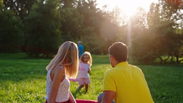 Gledelig barndom for en vakker liten jente og foreldrene sammen nyter den solfylte dagen i parken, mens den fryser på gresset. – stockvideo