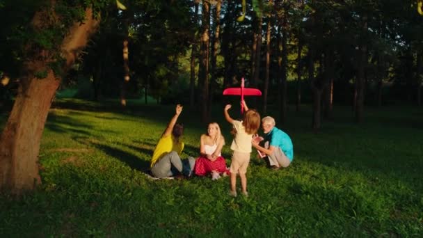 W zielonym parku charyzmatyczni rodzice z dziećmi i dziadkiem cieszą się razem z rodziną bawiącą się dużymi samolotami — Wideo stockowe