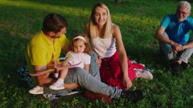Parkta büyük bir çimen tarlasında mutlu ve genç ebeveynler küçük tatlı kızlarıyla birlikte anın tadını çıkarıyorlar. Büyükbaba çimlerin üzerinde oturuyor.