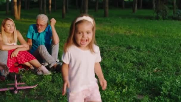 Στη φύση στο μεγάλο πεδίο της χλόης ευτυχισμένη και χαμογελώντας μεγάλο χαριτωμένο κορίτσι μπροστά από την κάμερα τρέχει και υπερβολική ταχύτητα καλή στιγμή με τους γονείς και τον παππού της — Αρχείο Βίντεο
