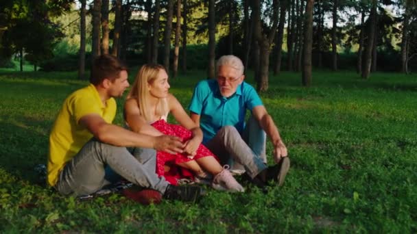 Im Park unten auf dem Gras glücklich junges Paar sehr attraktiv im Gespräch mit ihrem Vater alten Mann mit Brille sind sie glücklich, Zeit miteinander zu verbringen — Stockvideo
