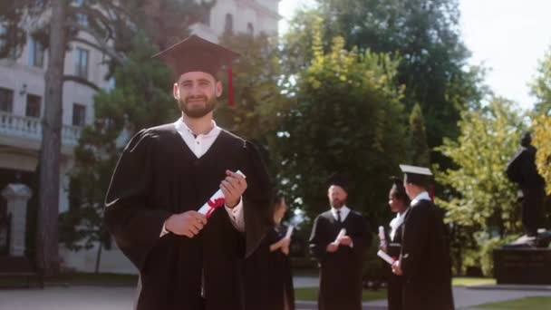 Перед камерою випускник хлопець з випускною шапкою позує перед камерою з дипломом в руках — стокове відео