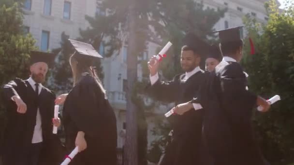 Estudiantes multiétnicos graduados bailando después de la graduación en el jardín de la universidad están muy felices y emocionados de tener diplomas en las manos — Vídeo de stock