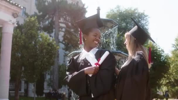Красивые многорасовые две студентки в выпускной день счастливо обнимают друг друга они носят выпускные костюмы и кепки — стоковое видео
