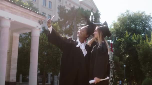 No jardim da faculdade moderna no dia da formatura na frente da câmera multirracial senhoras estudantes graduados posando animado tirando selfies com o smartphone — Vídeo de Stock