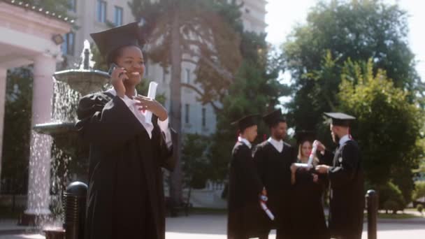 Charyzmatyczny jeden czarny pani student podekscytowany mówi przez telefon po ukończeniu studiów ona jest szczęśliwa inni studenci są na tle dyskusji wszyscy razem w ogrodzie college 'u — Wideo stockowe