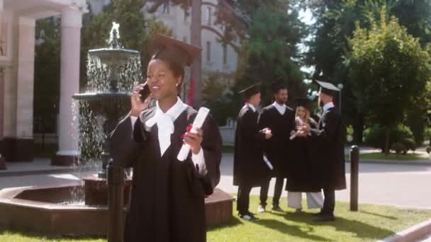 В день окончания школы в саду колледжа красивая молодая чернокожая женщина разговаривает по телефону после того, как она закончила другие студенты многорасовые обсуждения в группе на заднем плане — стоковое видео