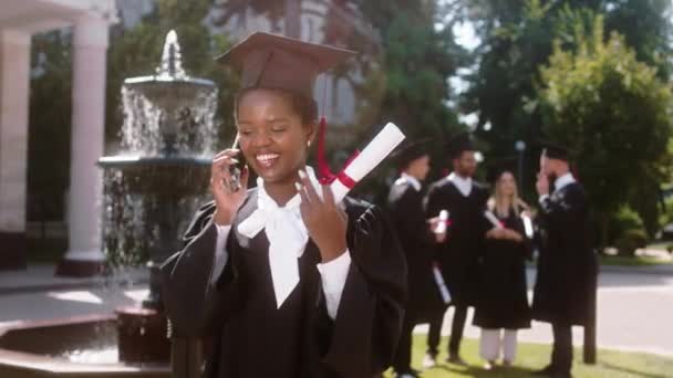 Señora negra muy hermosa en el día de la graduación tomar el teléfono y llamar a alguien que habla y sonriendo al mismo tiempo después de que se graduó en el fondo otros estudiantes discutiendo en grupo todos — Vídeo de stock