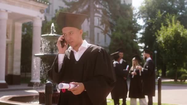 Estudante bonito depois que ele foi graduado chamado alguém e discutir muito animado enquanto segurando diploma no jardim da faculdade — Vídeo de Stock