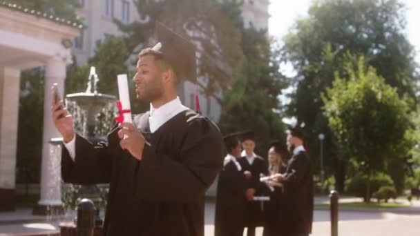Przystojny czarny facet po ukończeniu studiów weź telefon i zadzwoń do kogoś i mówi z aparatem pokazuje dyplom on jest bardzo podekscytowany tło inni studenci mówi na grupie — Wideo stockowe