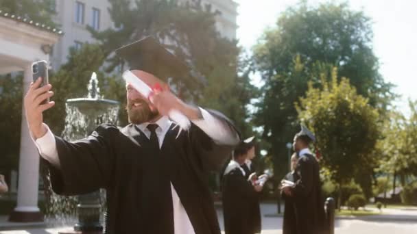 V den promoce vzrušený dospělý student absolvent pomocí smartphonu volat s fotoaparátem ukazuje svůj diplom a jeho skupina kolegů v zahradě vysoké školy — Stock video