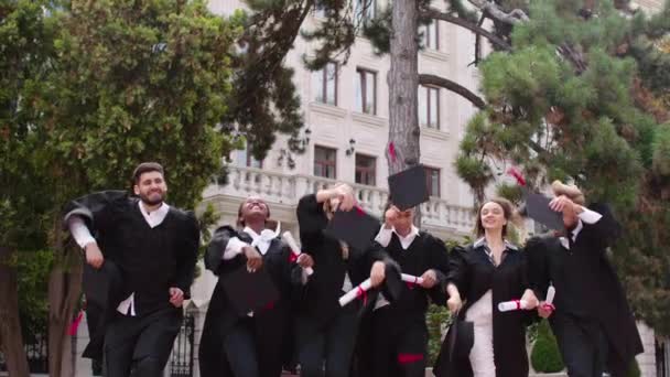 Возбужденные многорасовые студенты выпускников счастливо бросить выпускные шапки в саду колледжа они чувствуют себя отлично все вместе — стоковое видео