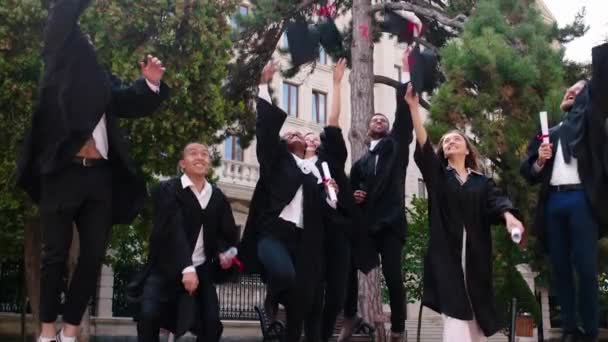 Studenti laureati multietnici nel giardino del college felici camminando tutti insieme davanti alla telecamera poi saltare e vomitare i berretti di laurea molto eccitati e in possesso di diplomi — Video Stock