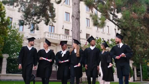 Grupo de estudantes carismáticos e bonitos graduados multirraciais no jardim da faculdade na frente da câmera andando todos os grupos juntos sorrindo grande discutir todos juntos vestindo bonés de formatura — Vídeo de Stock