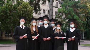 Coronavirus salgınında, üniversite parkında diplomaları elinde tutarak kameranın önünde poz veren çok ırklı mezunlar grubu.