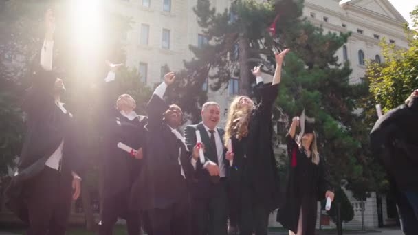 Opgewonden met een grote glimlach groep van multiraciale studenten afgestudeerden samen met de college directeur gooien de afstuderen caps ze knuffelen elkaar in de college tuin — Stockvideo