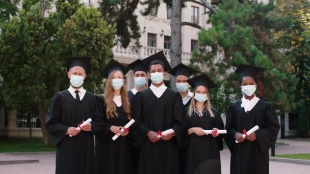 В пандемии Коронавируса группа многорасовых выпускников позирует перед камерой в защитной маске, держа дипломы в руках в парке колледжа — стоковое видео